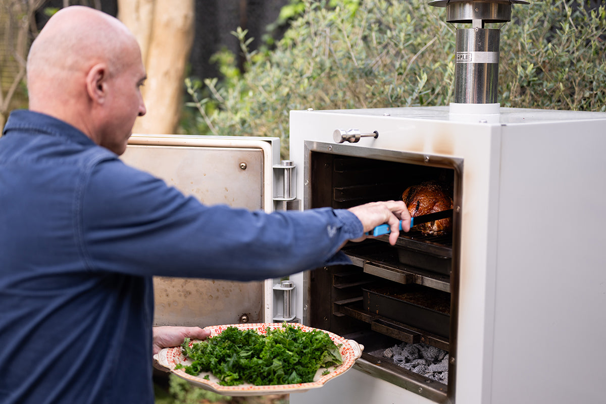 Man cooking a Chicken in an Charlie outdoor oven with a plate of greens nearby.