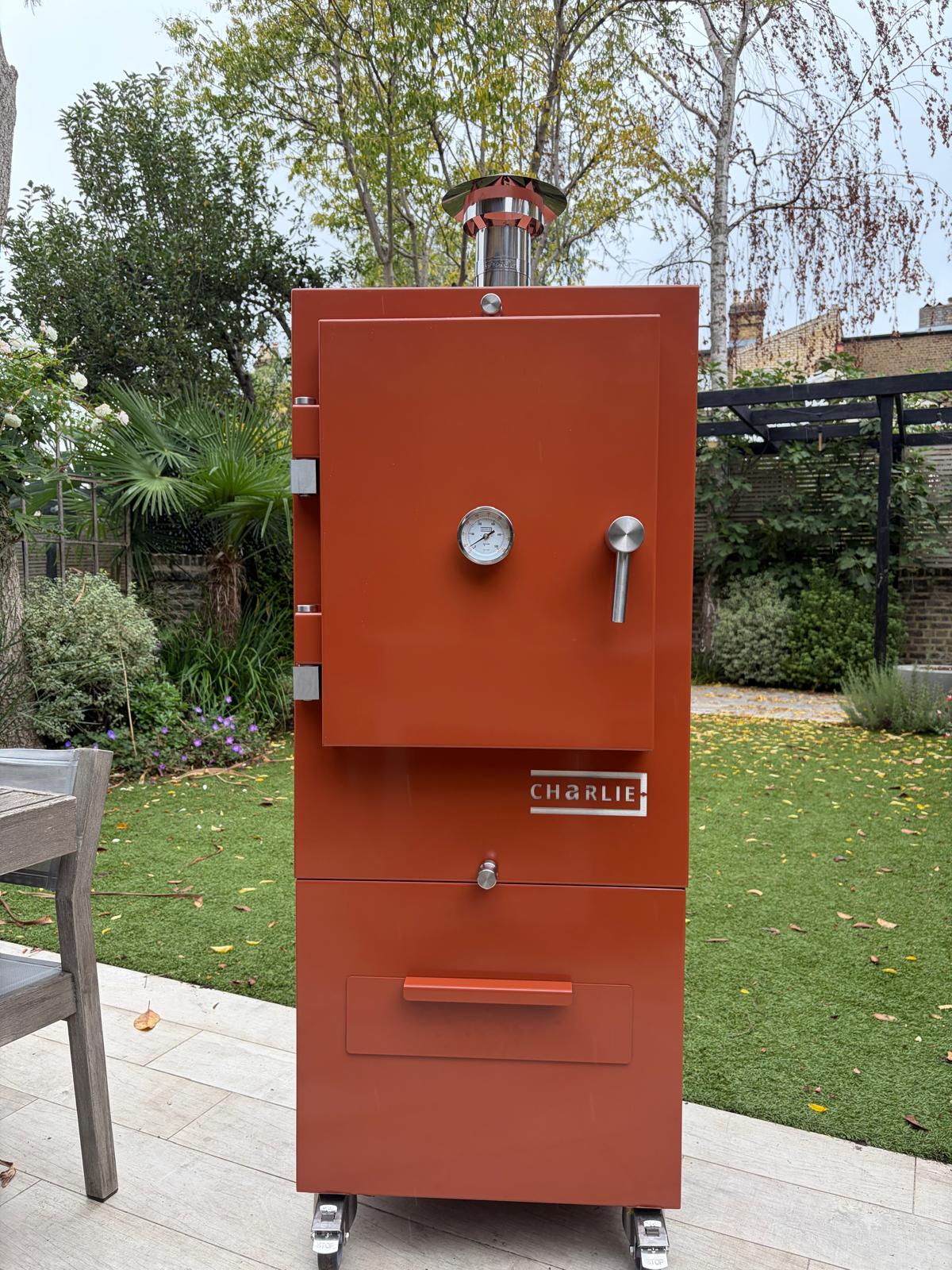 Charlie Charcoal Oven in cinnamon, a coppery orange-brown, freestanding on a garden patio, lifestyle view