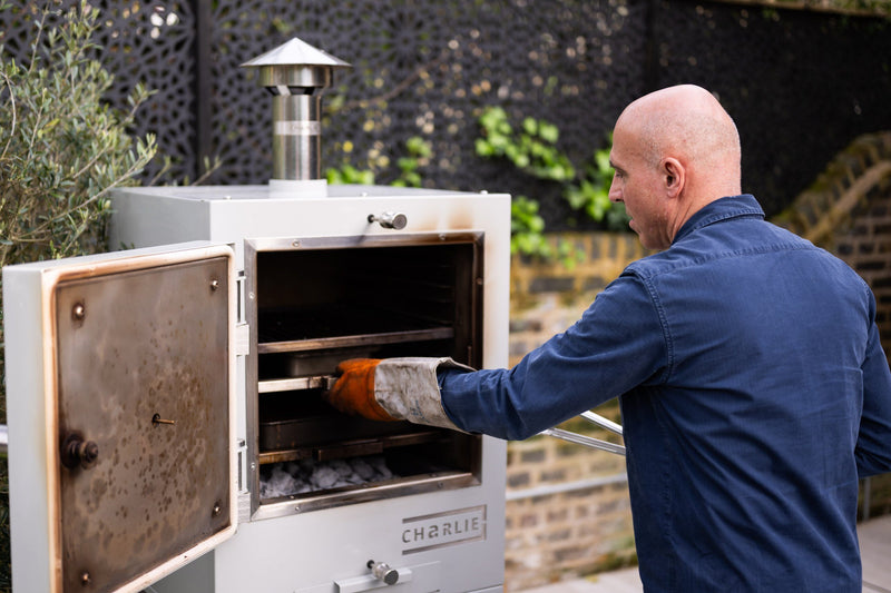 A cook in a heat glove sliding out a roasting tray from a Charlie charcoal oven