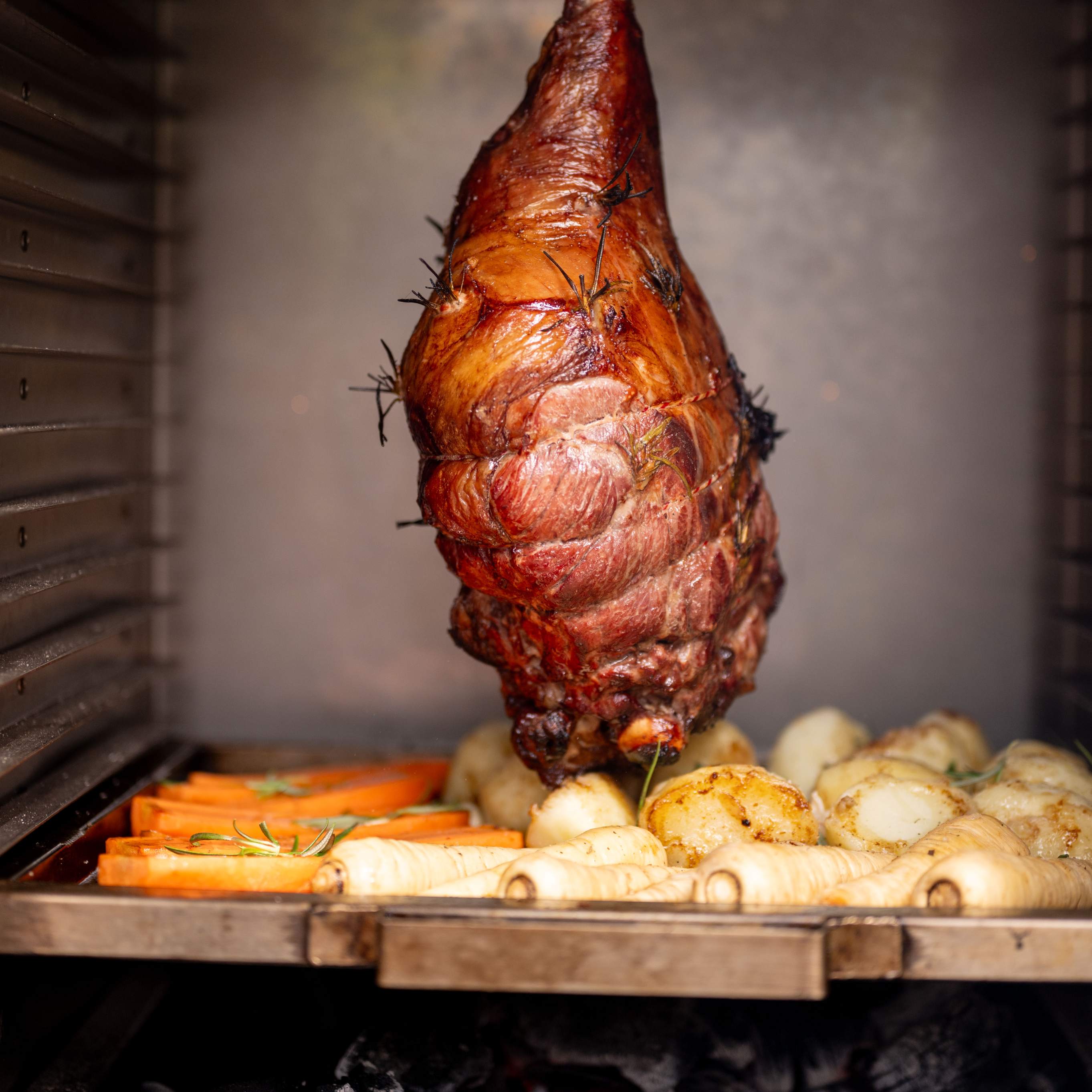 A rack of ham being roasted on a flat bed plancha rack inside an oven, surrounded by root vegetables.