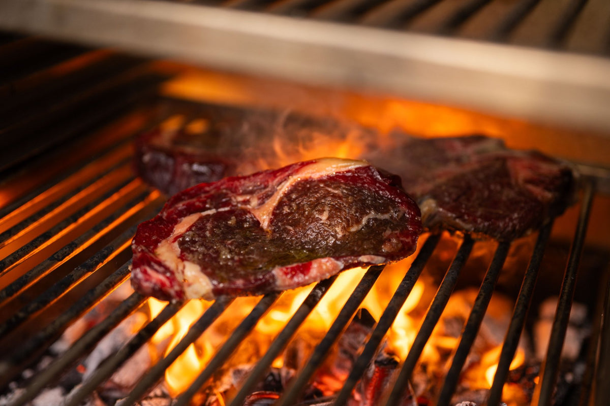 Ribeye steaks searing over glowing charcoal on the bottom grill rack of a Charlie Oven
