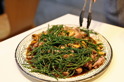 A plated dish featuring mussels, pieces of seared meat, and vibrant green samphire, all drizzled with a rich, dark sauce. A person is using tongs to serve the dish.
