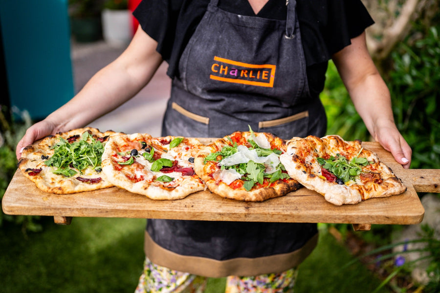 Four homemade pizzas topped with rocket and Parmesan on a long wooden board, held by a cook in a Charlie Oven apron