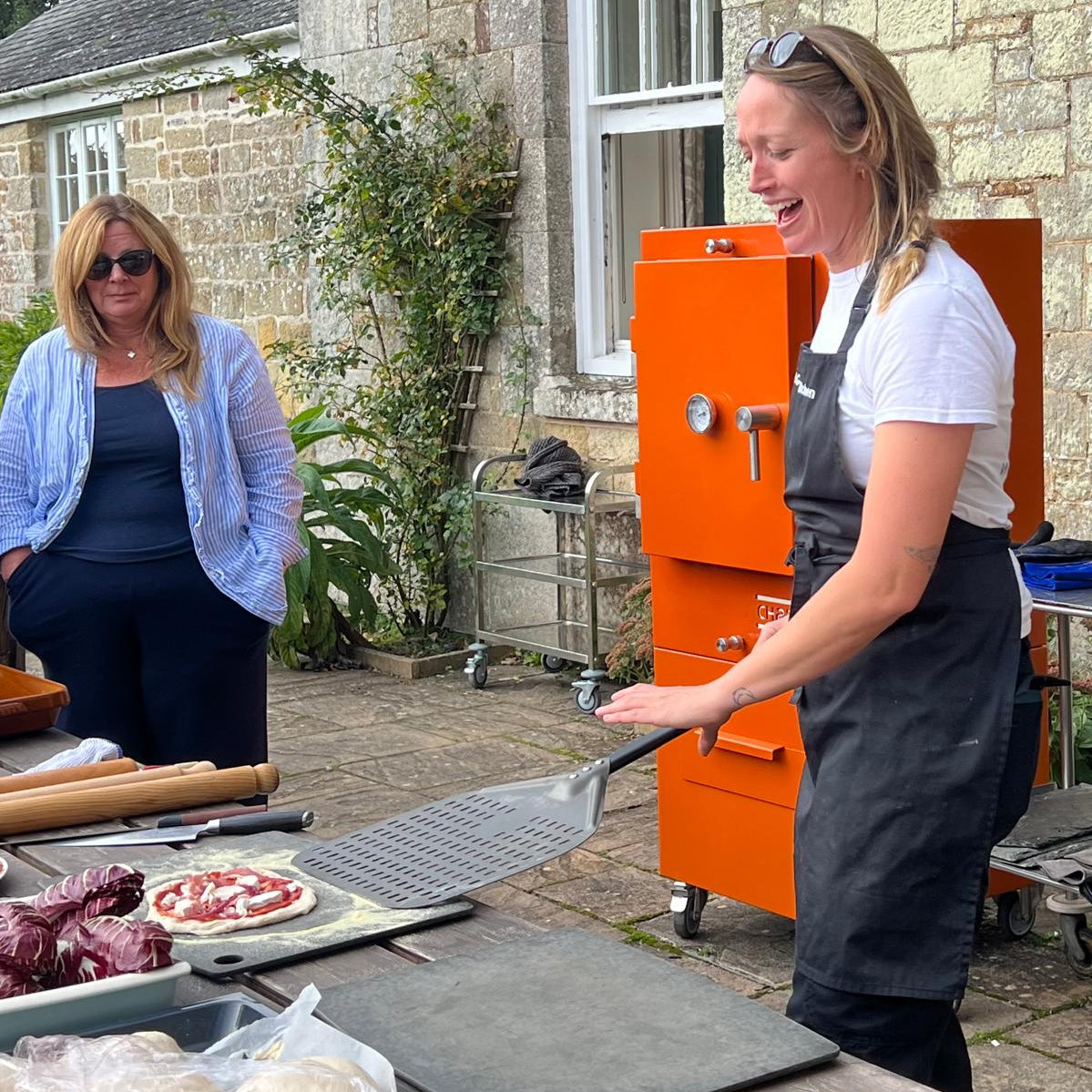 Two women outdoors, one preparing food in the charlie oven the other standing nearby.