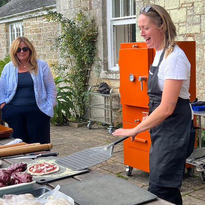 Two women outdoors, one preparing food in the charlie oven the other standing nearby.