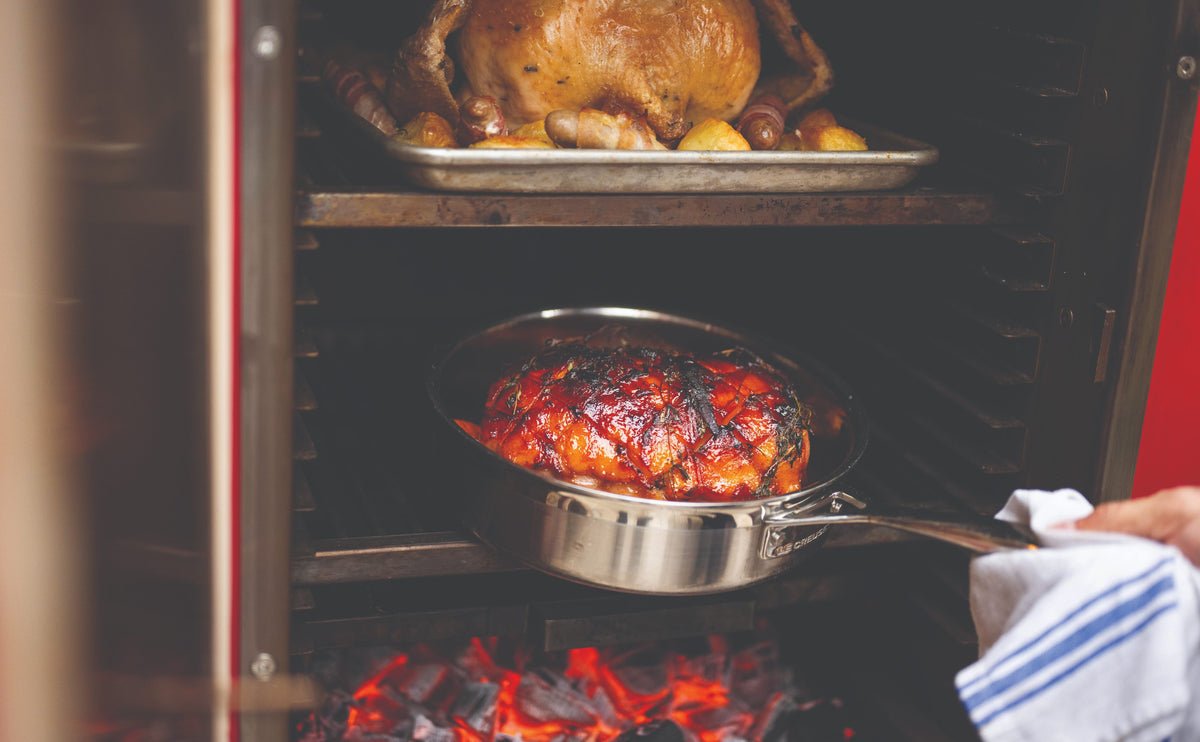 A Christmas turkey roasting on an upper level of a Charlie Oven, with a glazed ham over the glowing charcoal below
