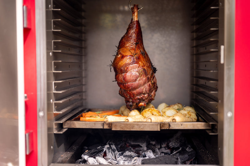 A leg of lamb hanging over a tray of roast potatoes inside a Charlie charcoal oven.