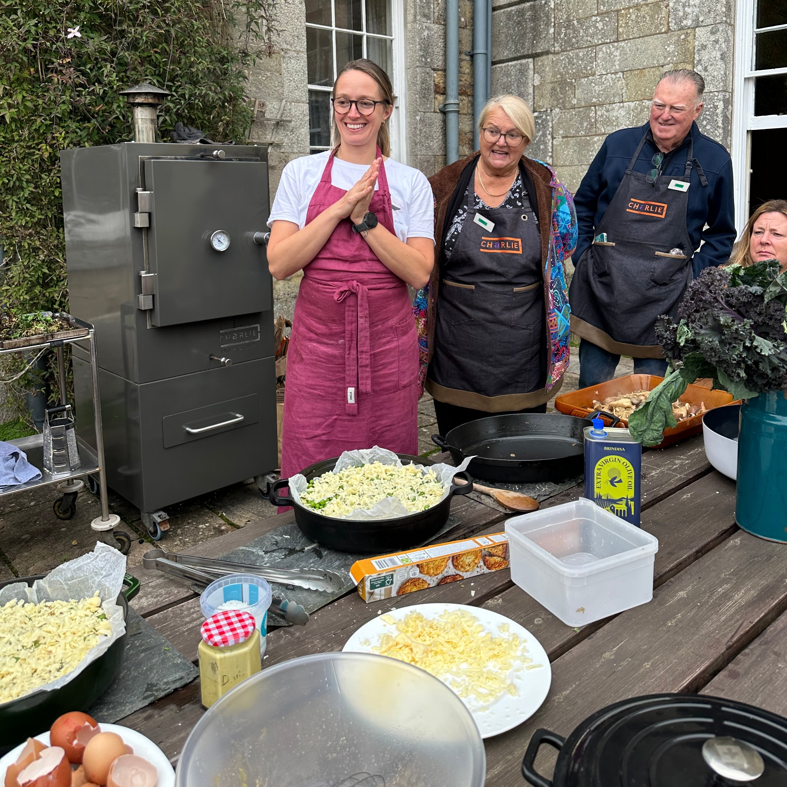 Three people standing outdoors near a Charlie grill with food and cooking ingredients.