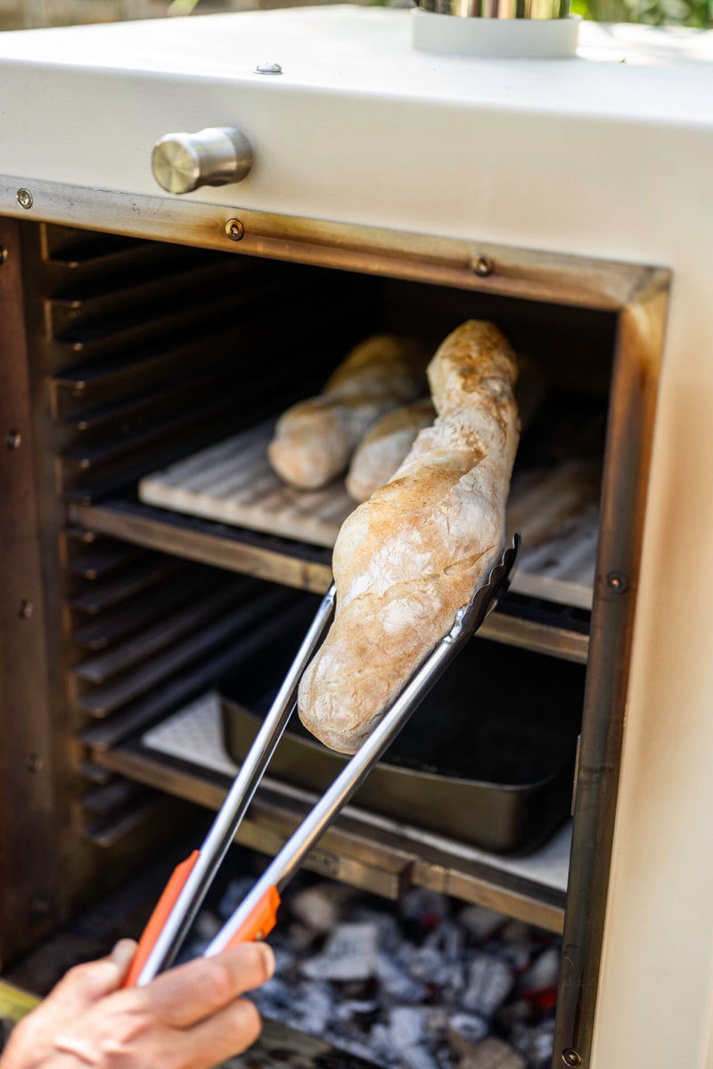 Lifting a baguette off the baking stone in a Charlie charcoal oven, with charcoal glowing on the grates below
