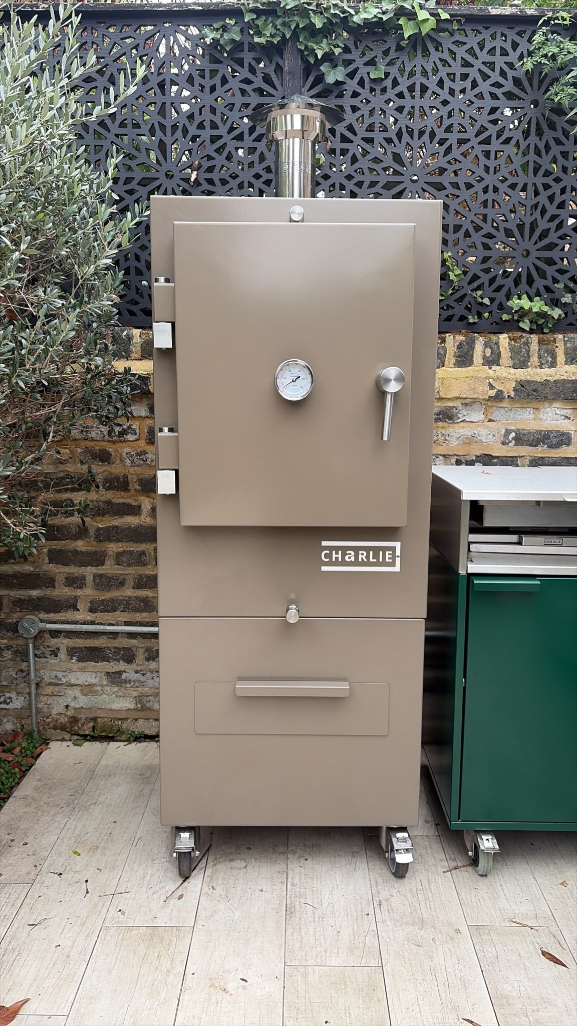 Charlie Charcoal Oven in nutmeg, a taupe brown, beside a green outdoor kitchen cabinet against a brick garden wall, lifestyle view 