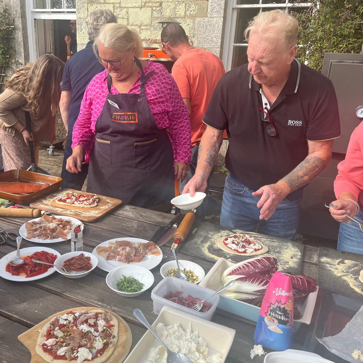 People preparing pizzas outdoors at a table with ingredients and utensils.