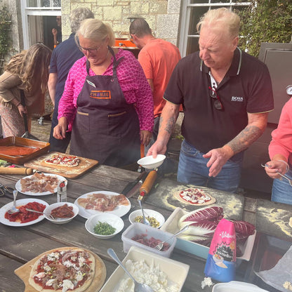 People preparing pizzas outdoors at a table with ingredients and utensils.
