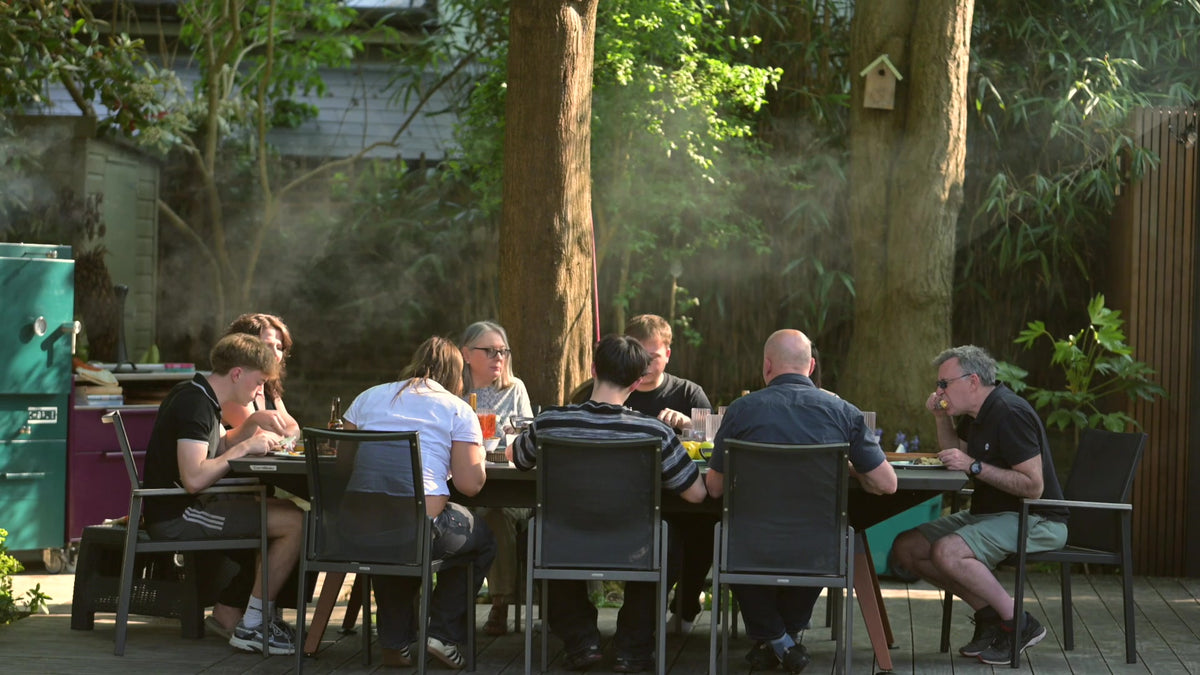 A long garden table laid for dinner with a Charlie Oven smoking behind