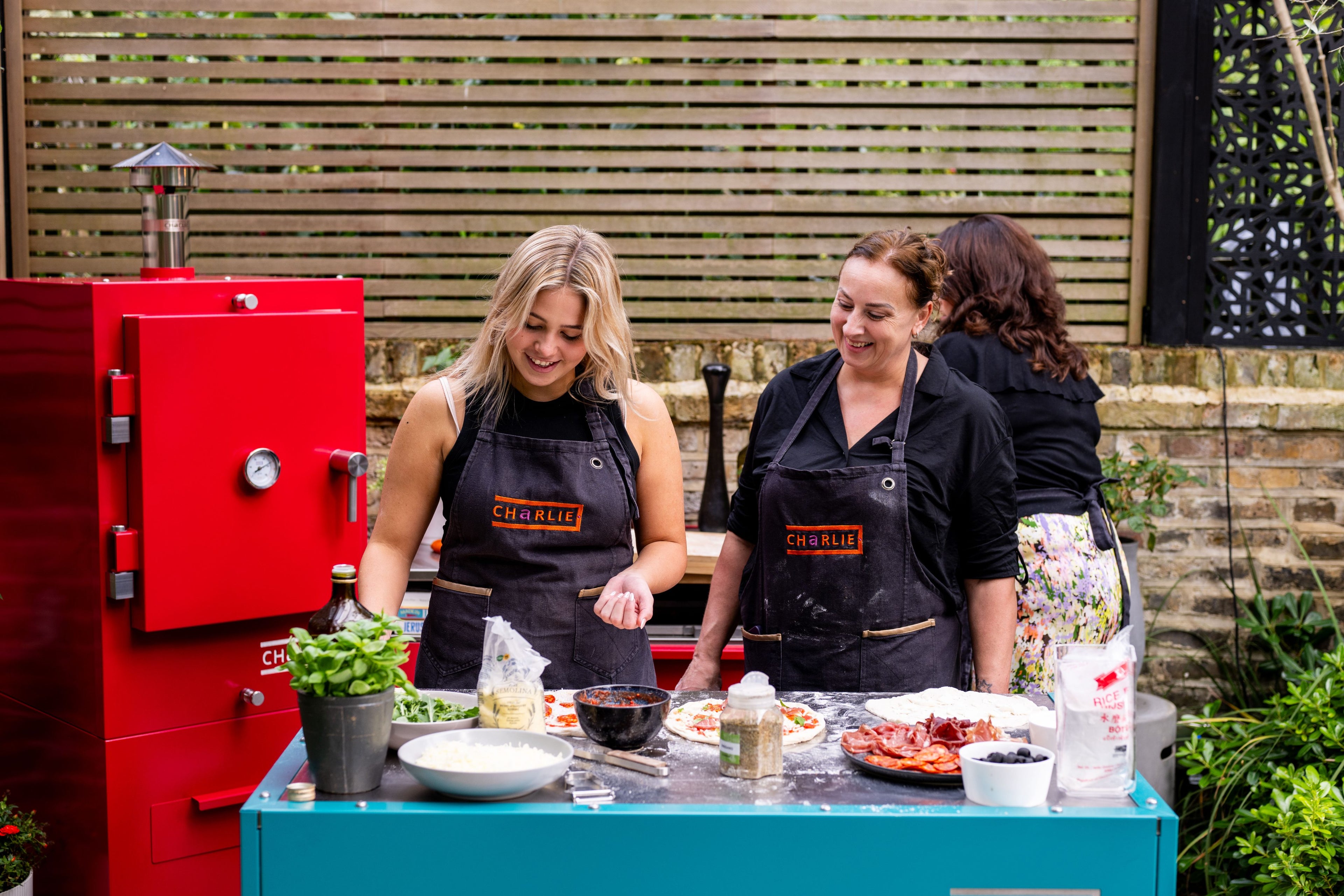 Two women in black aprons preparing food outdoors next to a red Charlie Oven.