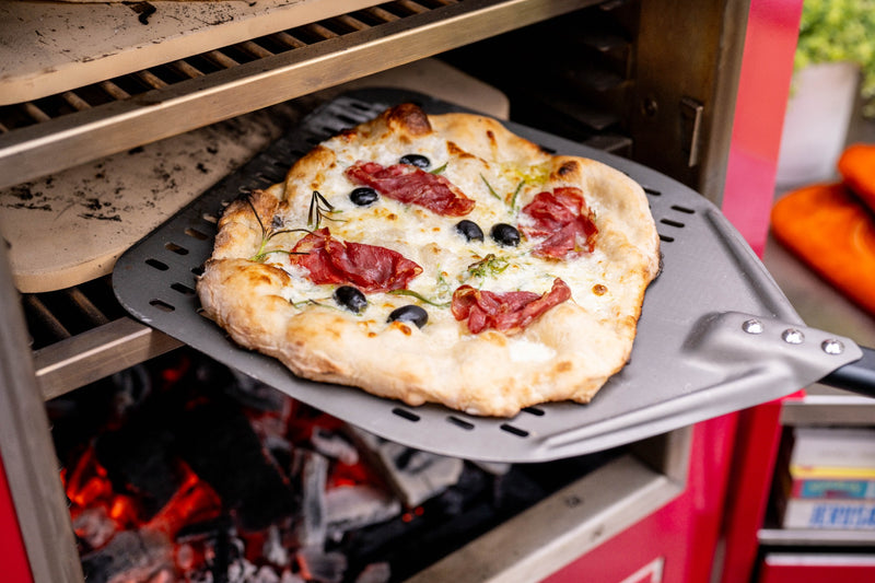 A pizza coming out of a Charlie charcoal oven on a peel.
