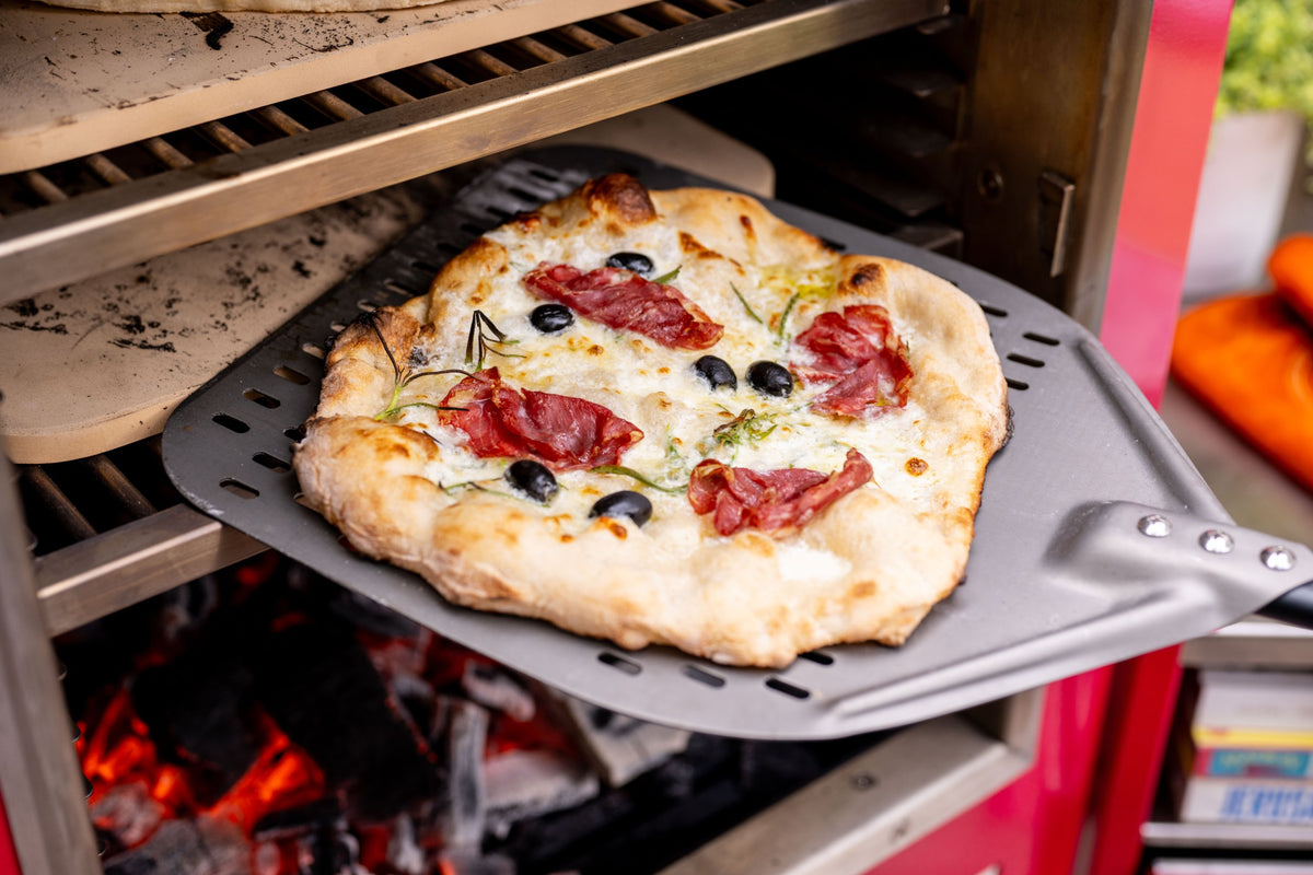 A pizza coming out of a Charlie Oven on a perforated peel, with pizza stones on the rack levels above and charcoal glowing below