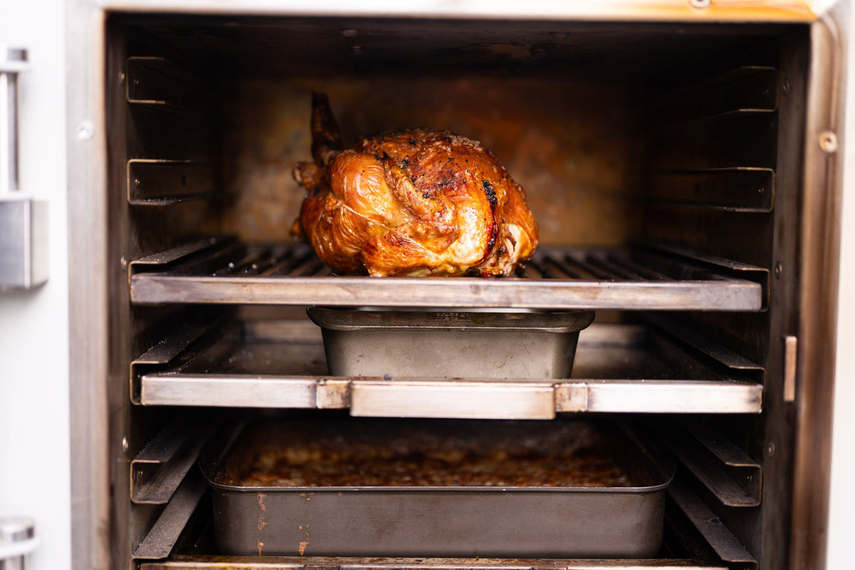 A roast chicken on the top rack with roasting tins cooking on the levels below