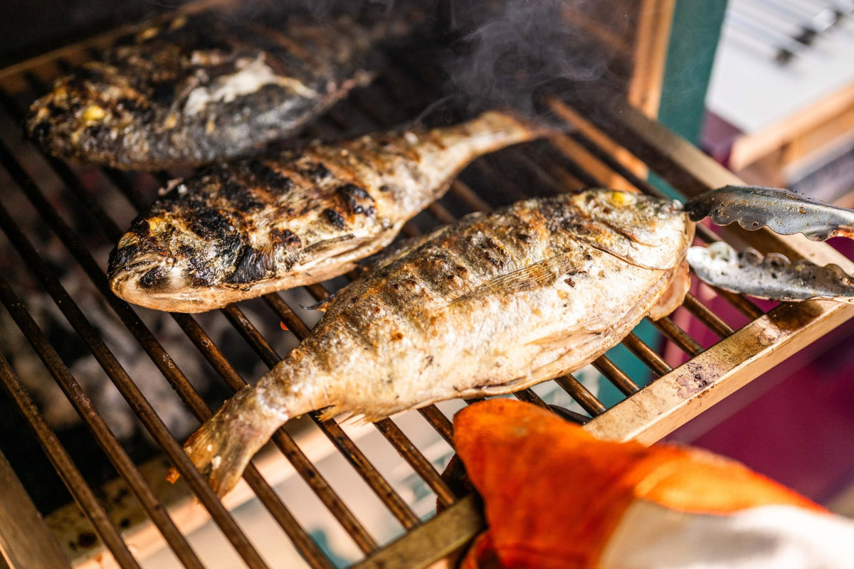 Three whole sea bream with charred skin, turned with tongs on the grill rack of a Charlie Oven